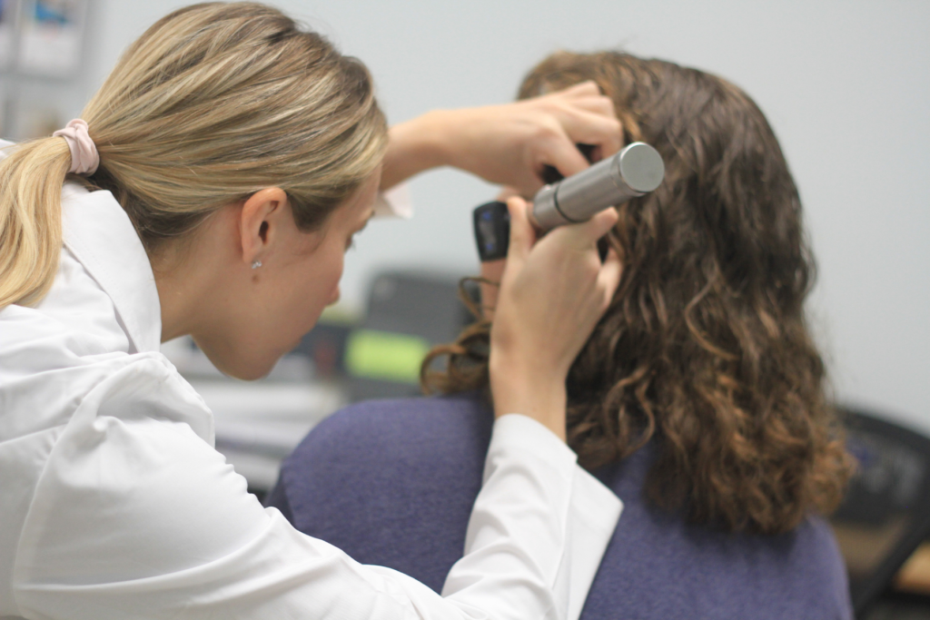 blonde audiologist with an NPI in white coat using an otoscope to view a patient's ear